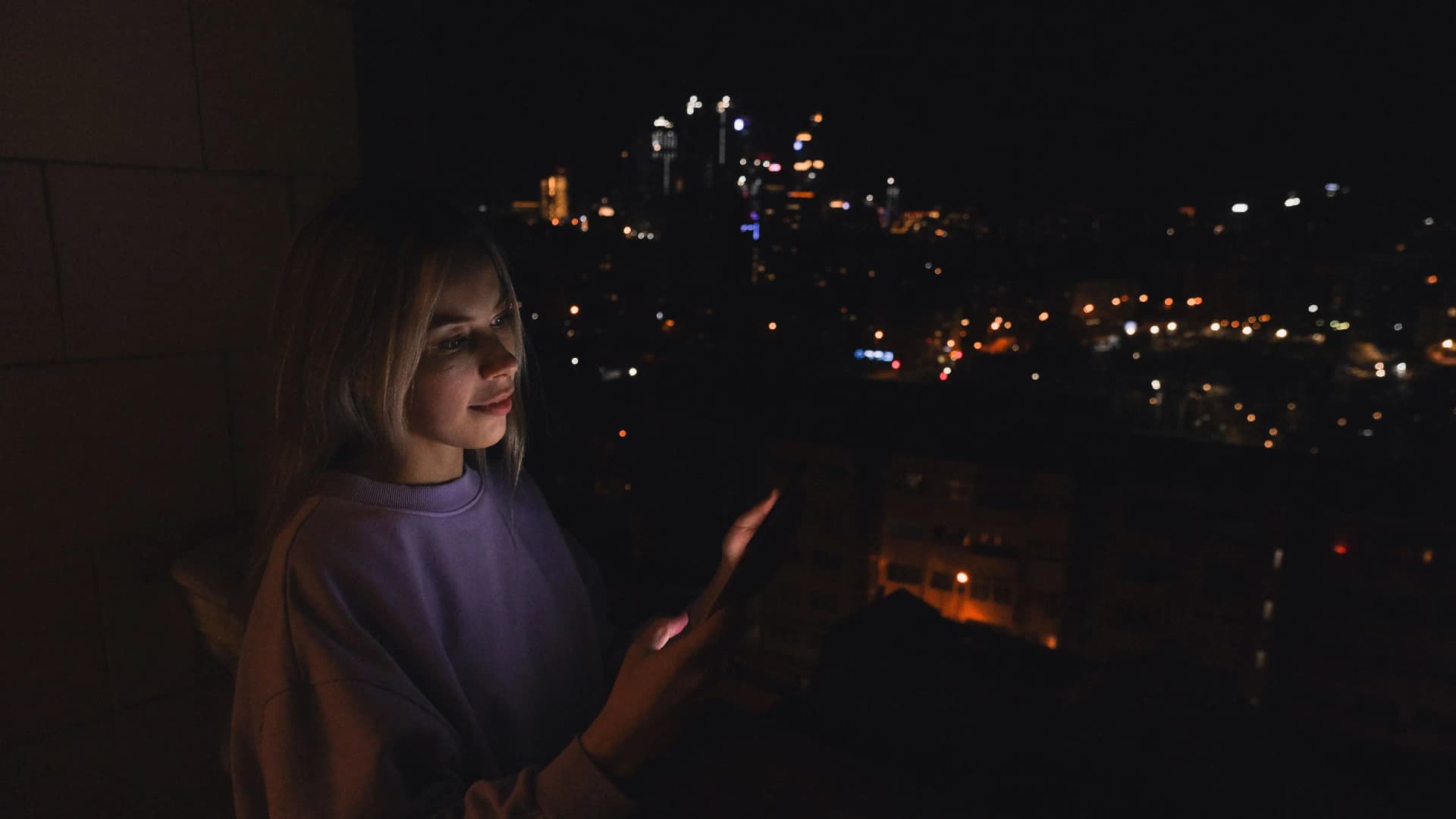Alguien en un balcón de noche, iluminado por su móvil, con la ciudad debajo