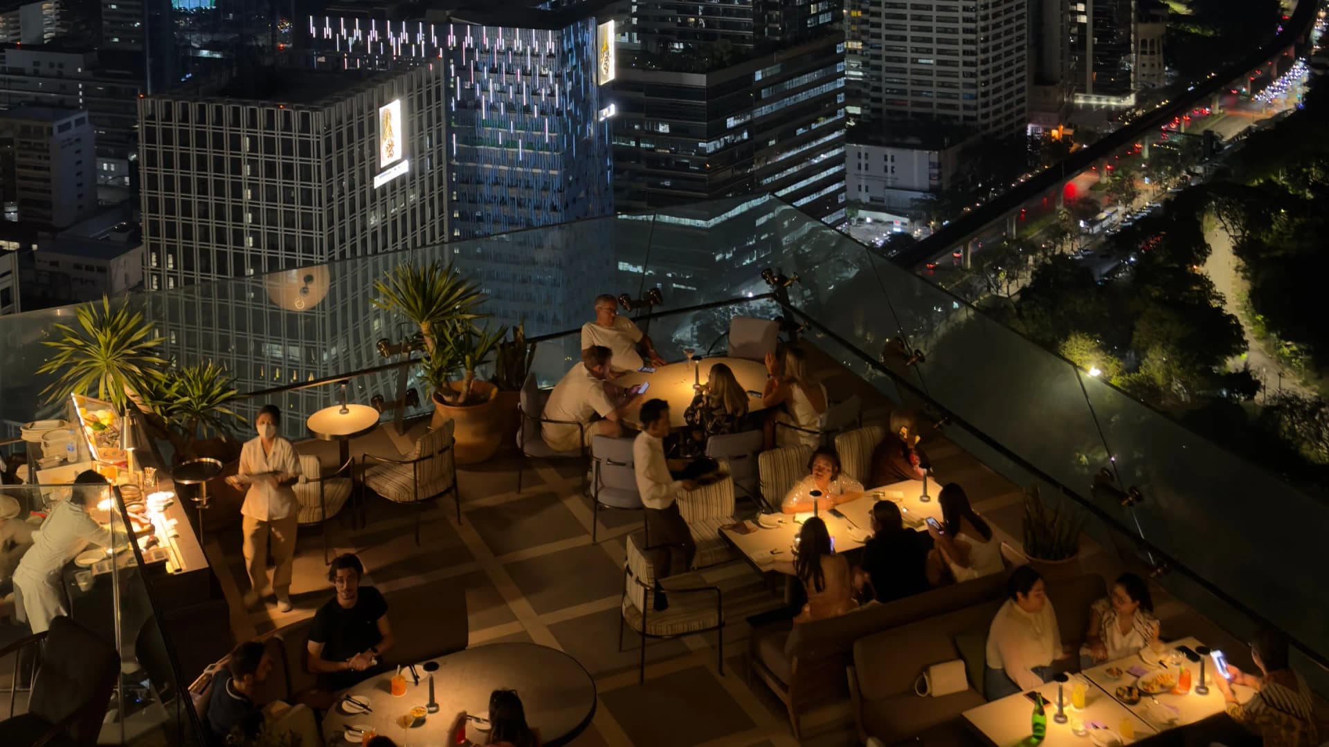 Terraza en altura de un local por la noche, con clientes en las mesas y la ciudad debajo